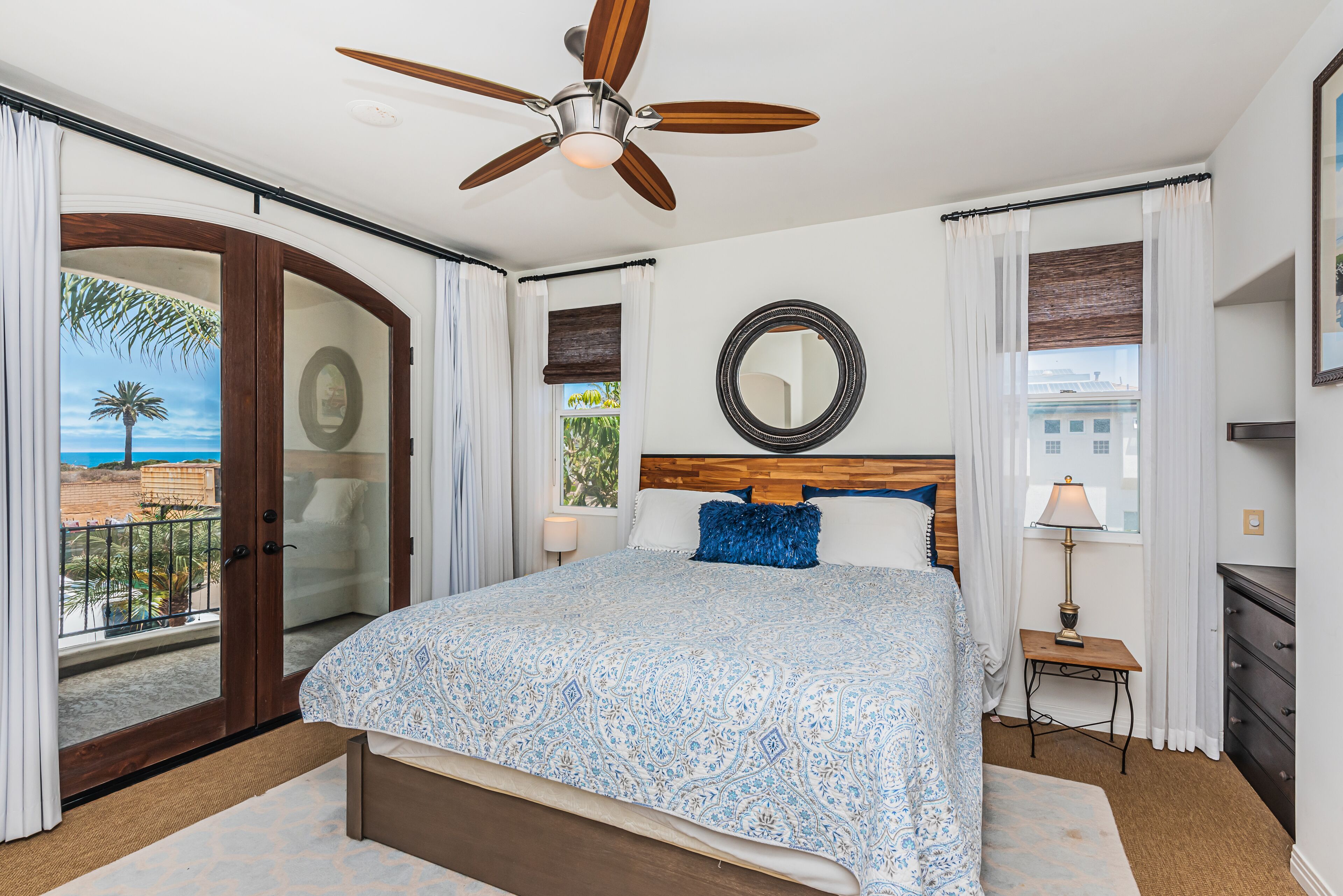 A bedroom with a large bed, a ceiling fan, a wooden headboard, a bedside table with a lamp, and a view of the outdoors.