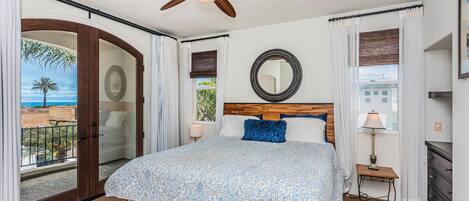 A bedroom with a large bed, a ceiling fan, a wooden headboard, a bedside table with a lamp, and a view of the outdoors.