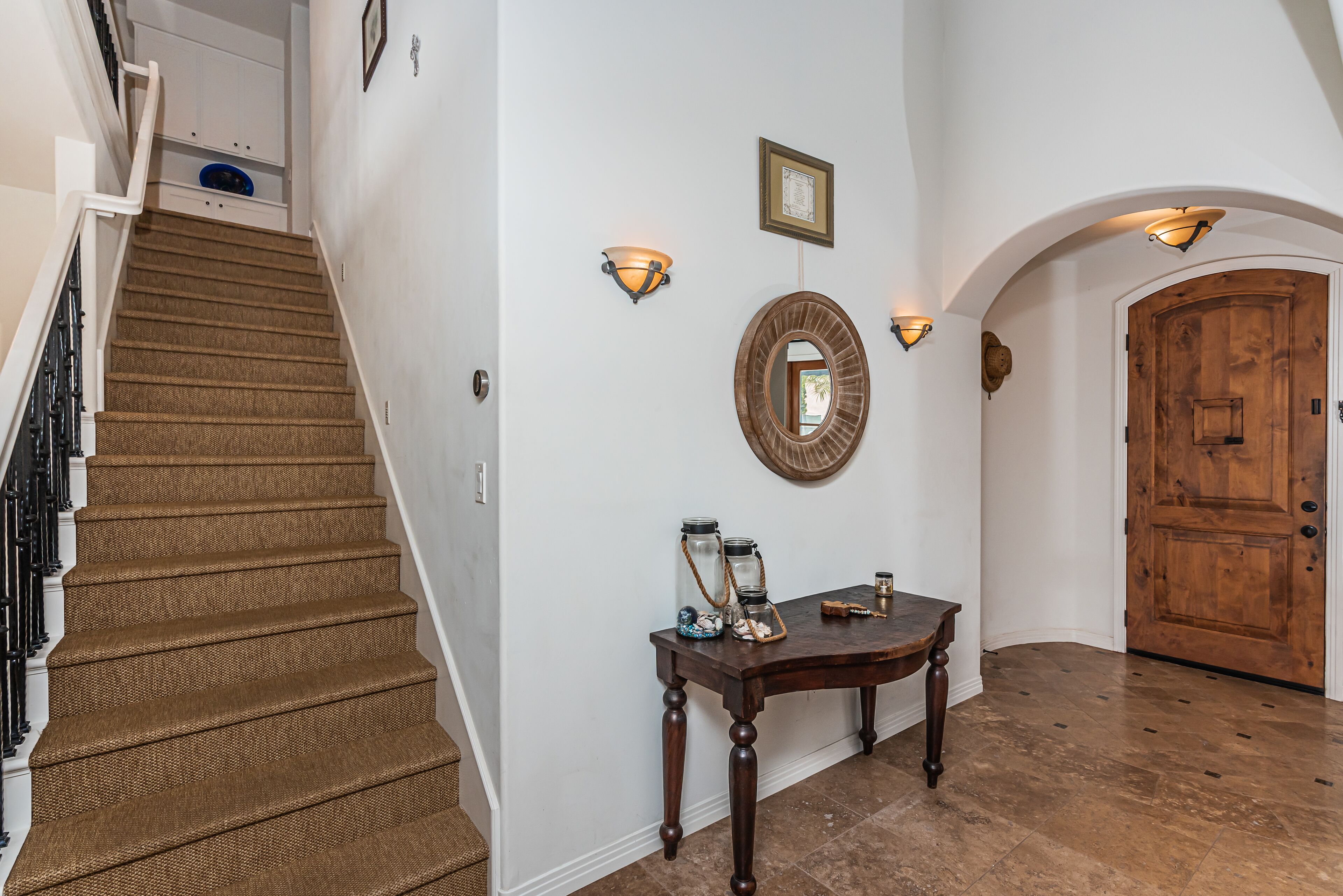 A staircase with a textured carpet, a wooden table with decorative items, wall sconces, and a wooden door.