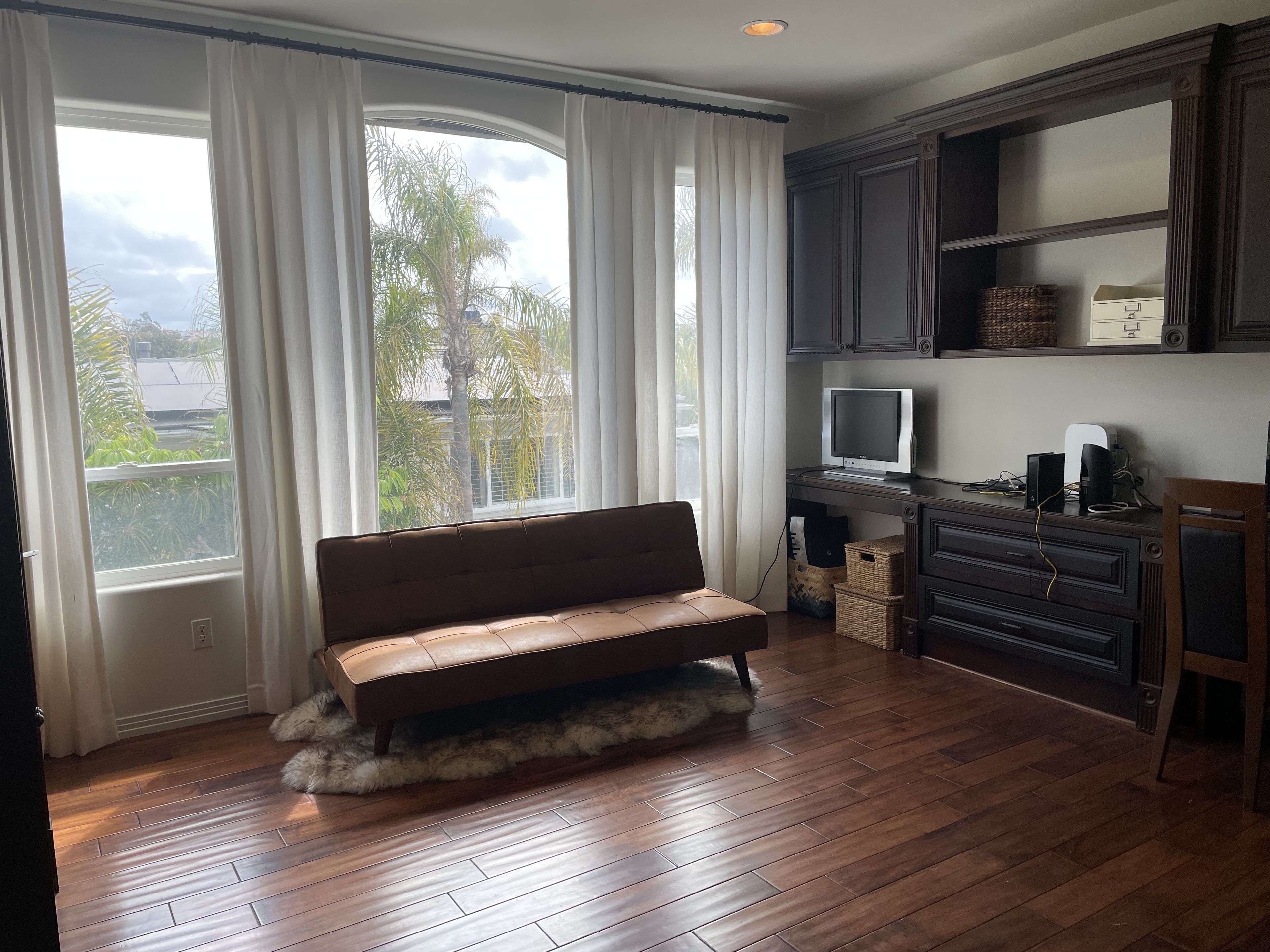 Una habitación con un sofá de cuero marrón, un escritorio de madera con una computadora y un monitor, y vistas a palmeras a través de amplios ventanales.