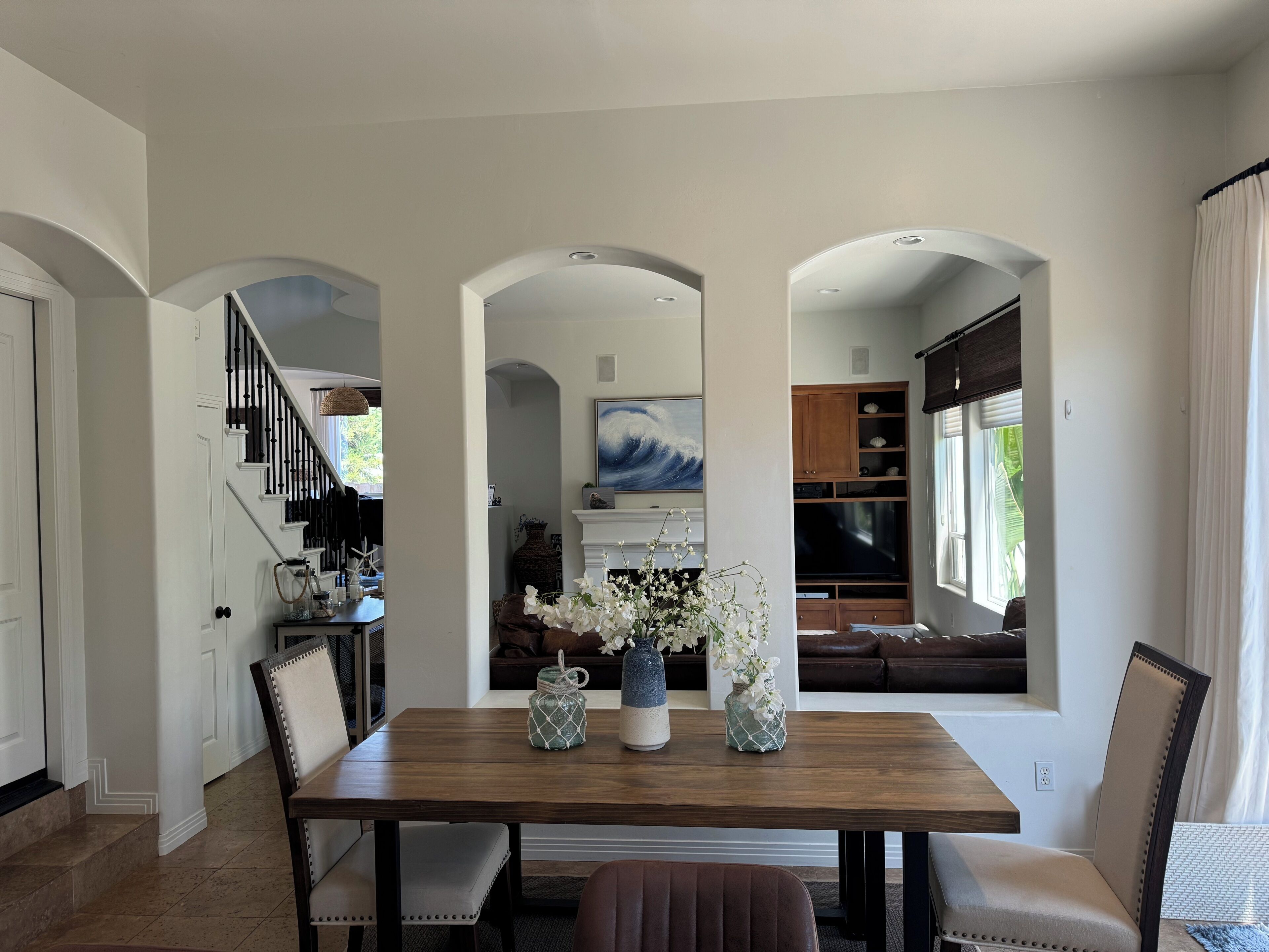A dining area with a wooden table and chairs, a vase with flowers, and a view of a kitchen and living area through an archway.