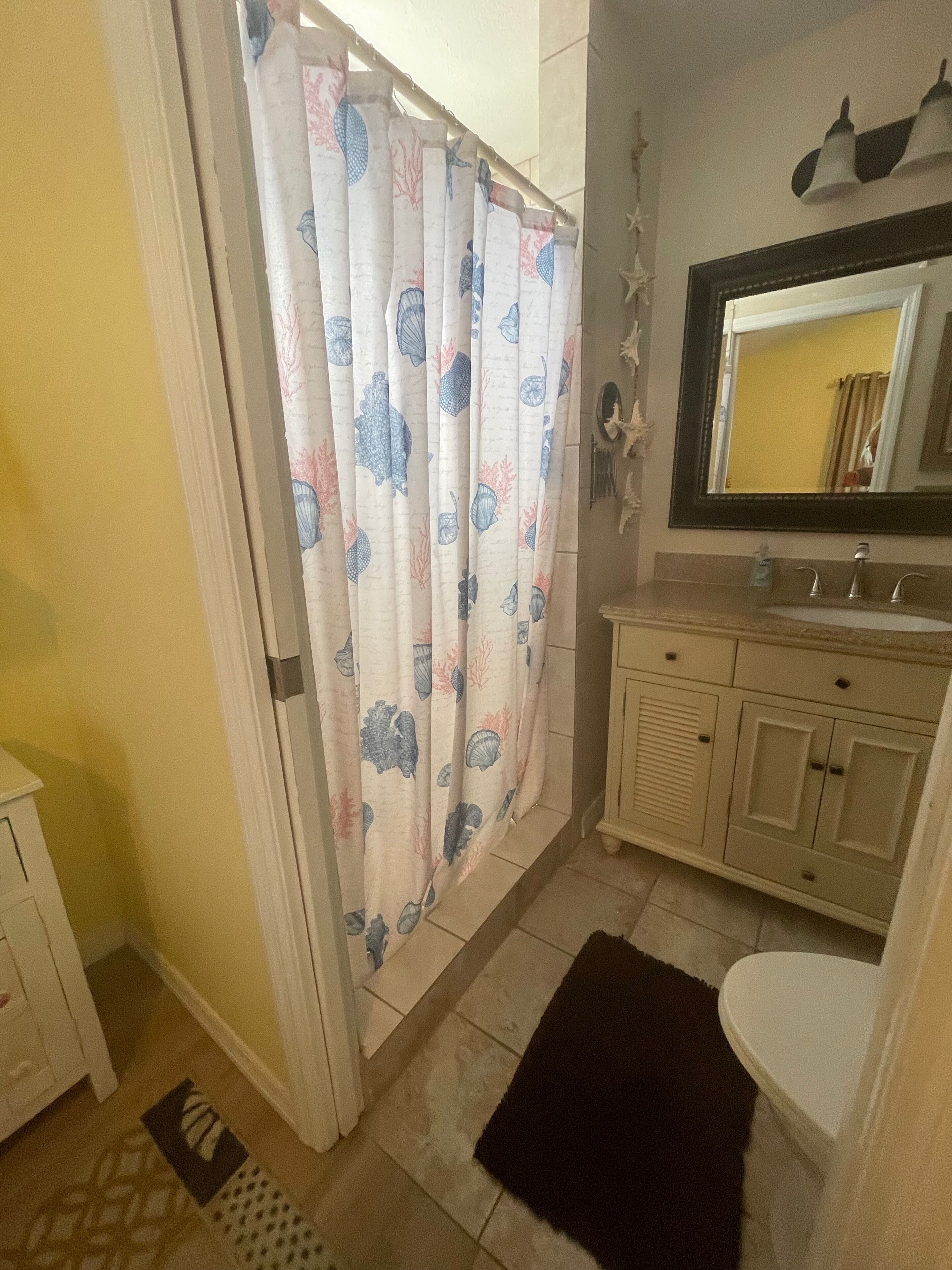 A bathroom with a shower curtain featuring a shell pattern, a sink with a mirror above it, and a small rug on the floor.