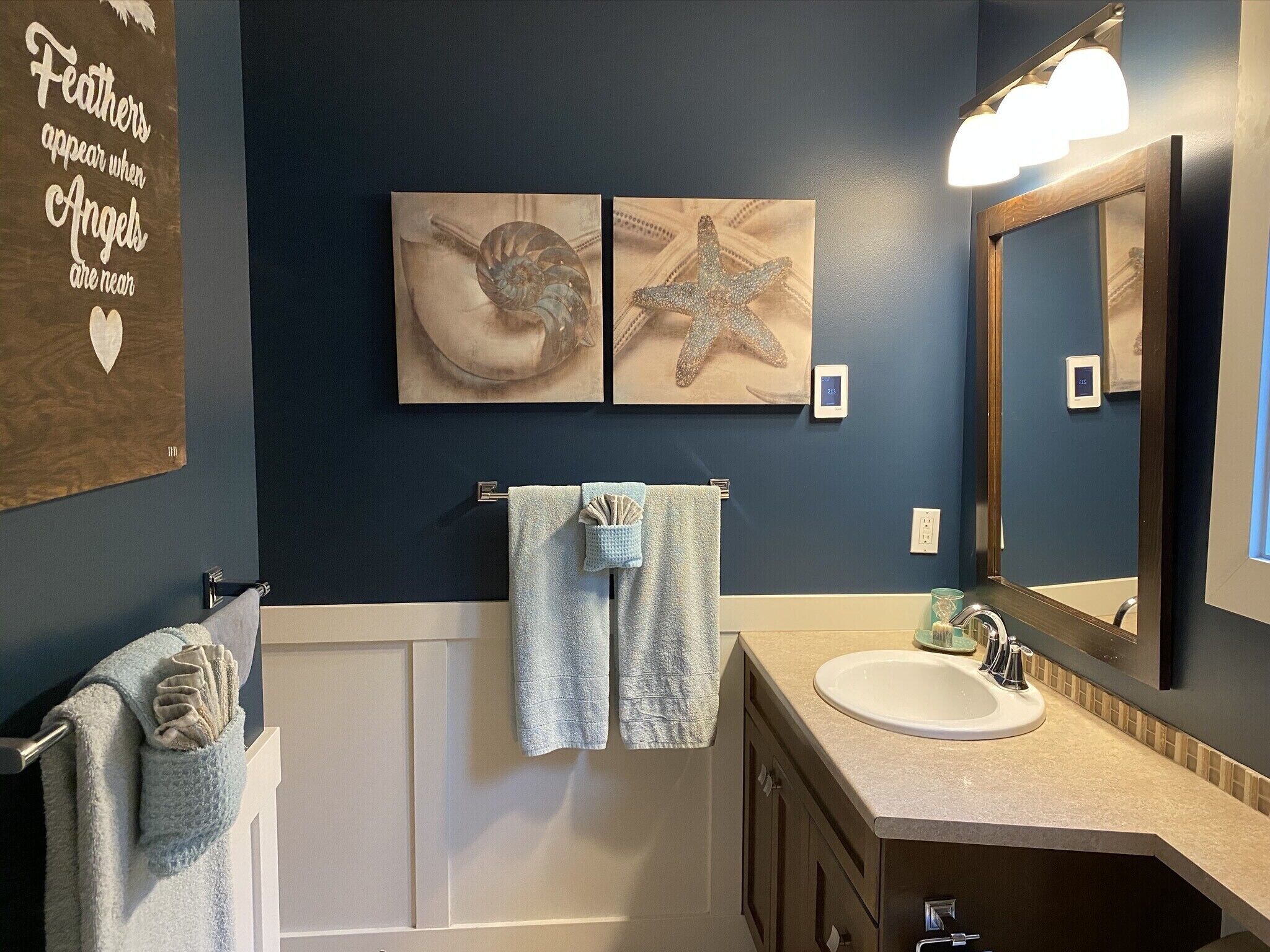 A bathroom with a dark blue wall, a white bathtub, a sink with a mirror, and two framed artworks depicting a seashell and a starfish.