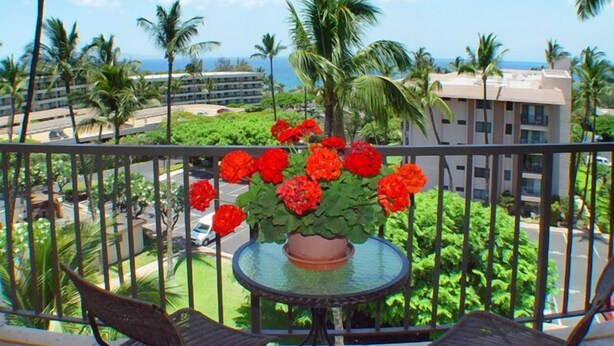 Ein Balkon mit einem runden Glastisch, roten Blumen und Blick auf Palmen und ein Gebäude.