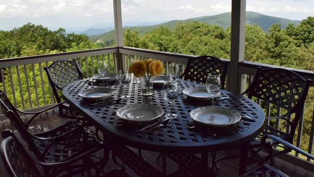 Una mesa redonda dispuesta para comer, con sillas, un florero con flores amarillas y una vista panorámica de montañas y árboles.