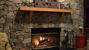 Una chimenea de piedra con fuego, un estante de madera encima y un sillón estampado con motivos florales al frente.