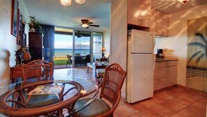 A spacious room with a dining area, kitchen, and a large window offering a view of the ocean. - Pikake - West Maui Beachfront Condo! 20 Feet from the Ocean! (Lahaina)