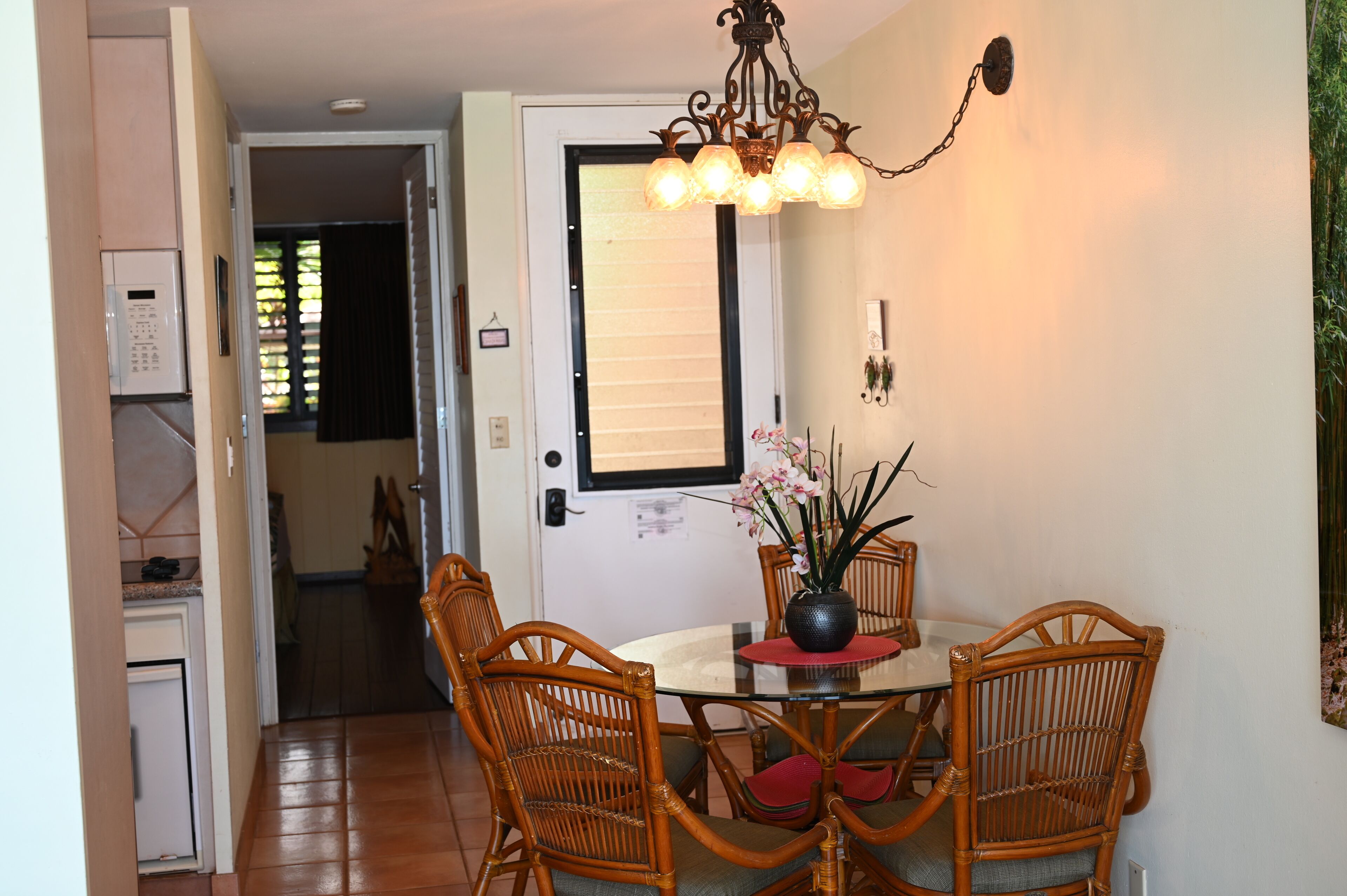 Un comedor con una mesa redonda, cuatro sillas de mimbre, un jarrón con flores y un aplique de pared.