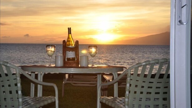 A table with two glasses and a bottle, set against a sunset view. - Pikake - West Maui Beachfront Condo! 20 Feet from the Ocean! (Lahaina)