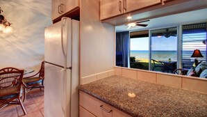 A kitchen with a refrigerator, wooden cabinets, a granite countertop, and a view of the ocean through large windows. - Pikake - West Maui Beachfront Condo! 20 Feet from the Ocean! (Lahaina)