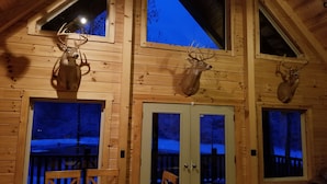 Interior de uma cabana de madeira com dois pares de chifres de veado nas paredes, uma mesa de jantar com cadeiras e um sofá.