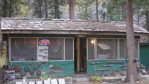 A rustic cabin with a green exterior, a neon 'OPEN' sign, and various tools displayed outside.