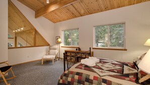 Une chambre avec un plafond en bois, un lit recouvert d’une courtepointe, deux fenêtres, une chaise et un bureau.
