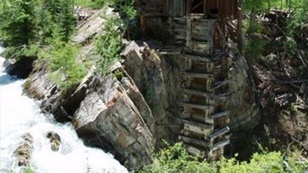 A rustic wooden structure built on a rock ledge by a river, surrounded by lush greenery and mountains in the background.