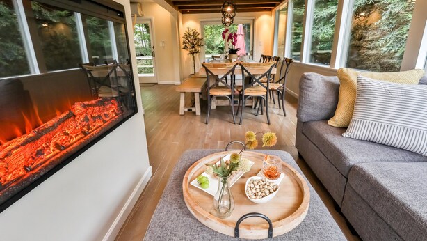 Una sala de estar acogedora con chimenea, un comedor con mesa y sillas de madera, y un sofá gris con cojines estampados.