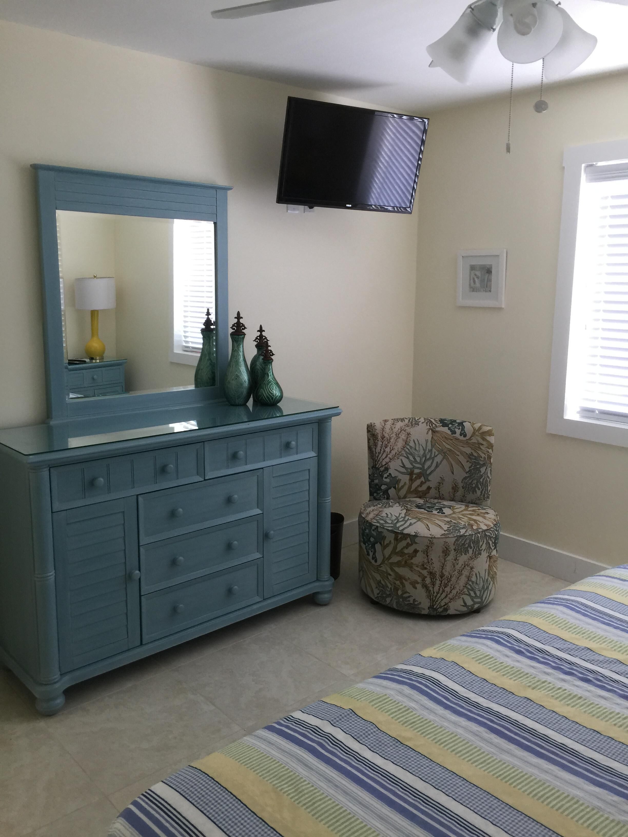 A bedroom with a blue dresser, a floral armchair, a bed with striped bedding, a flat-screen TV, and a ceiling fan.