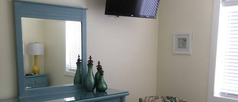 A bedroom with a blue dresser, a floral armchair, a bed with striped bedding, a flat-screen TV, and a ceiling fan.
