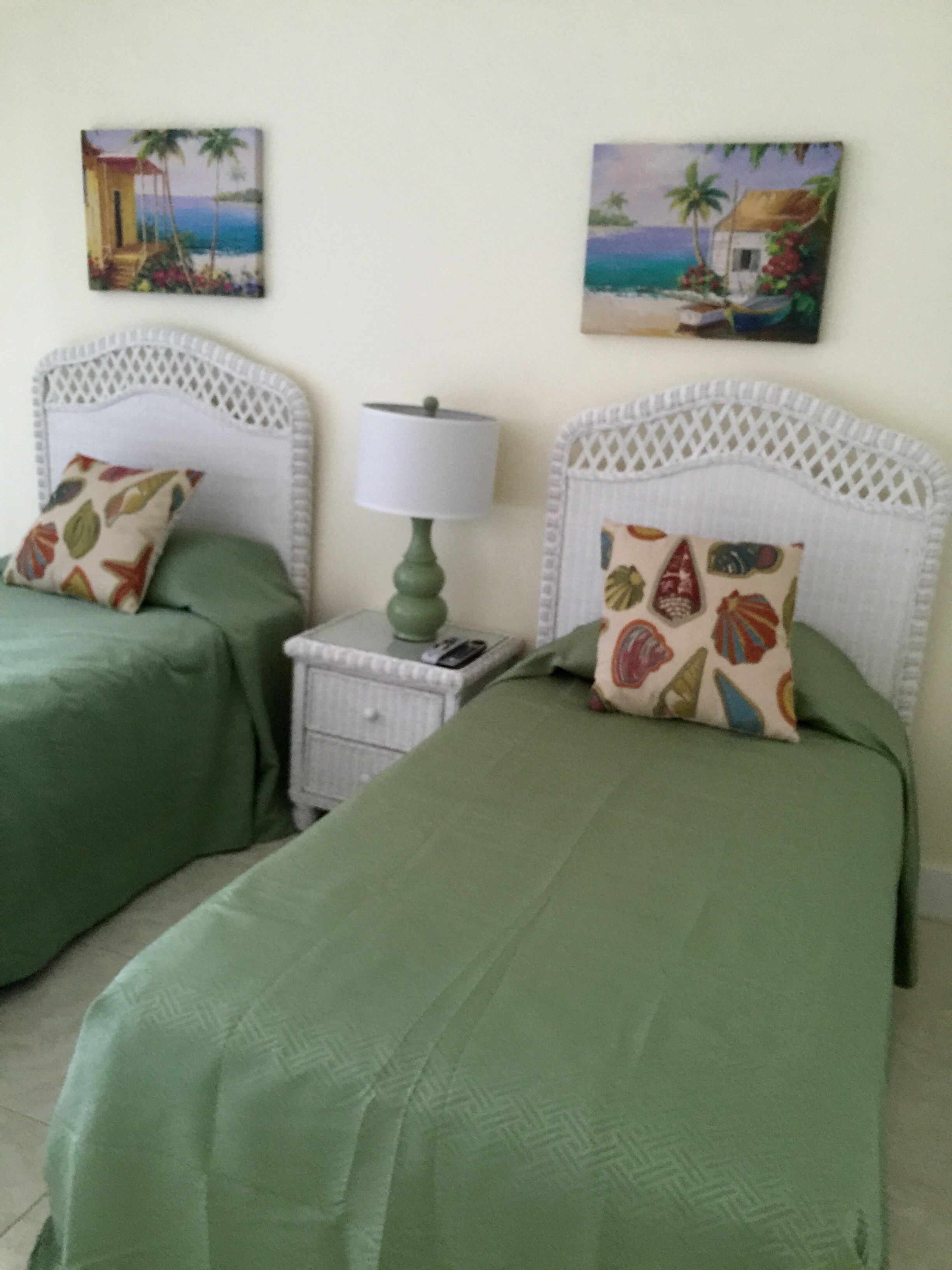 A room with two beds, each with a green bedspread and decorative pillows. There is a nightstand with a lamp between the beds.