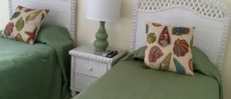 A room with two beds, each with a green bedspread and decorative pillows. There is a nightstand with a lamp between the beds.