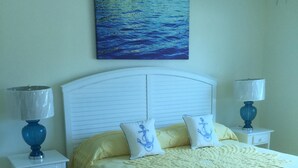 Un letto rifatto con cura, con una coperta gialla, due cuscini coordinati e una testiera bianca. Sono presenti due comodini con lampade, uno per lato del letto. Sopra il letto, un quadro raffigura un mare blu.