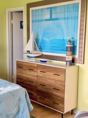 A bedroom with a wooden dresser, a bed with a blue bedspread, a painting of a lighthouse and sailboat on the wall, and a door with a blue sign.