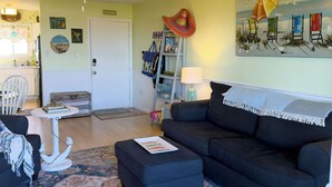 A living room with a dark sofa, a patterned rug, a ceiling fan, and a painting of beach chairs.