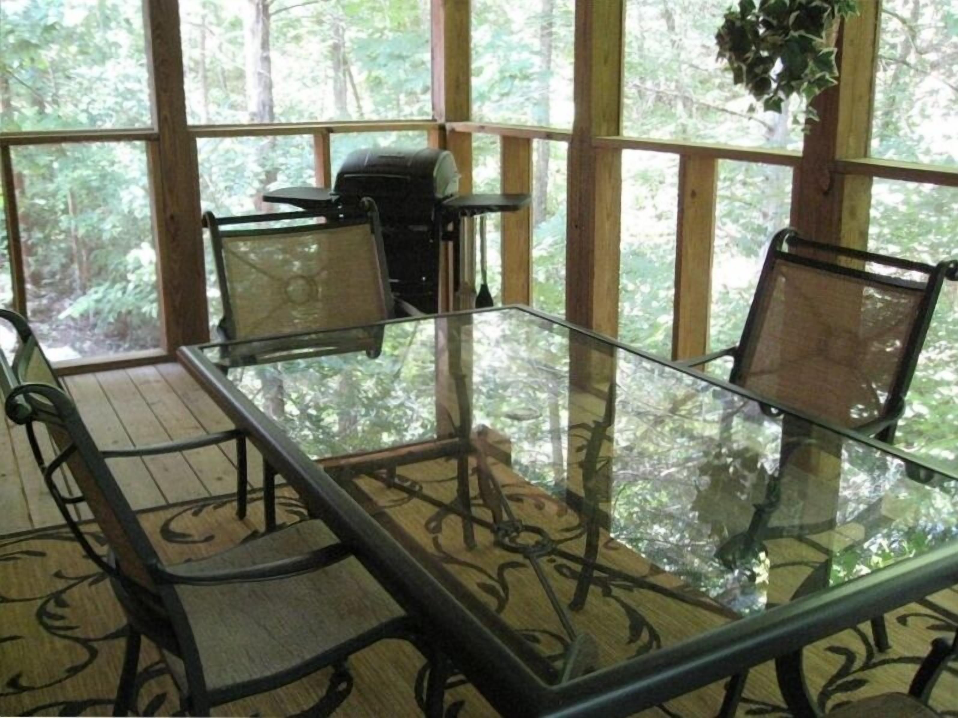 A glass-top table with four chairs, surrounded by wooden beams and large windows offering a forest view.
