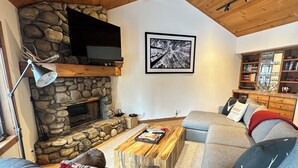 A cozy living room with a stone fireplace, a flat-screen TV mounted above it, and a large framed picture on the wall. - Exclusive Teton Pines Home off Teton Village Rd. 5 min from Teton Village Skiing (Wilson)