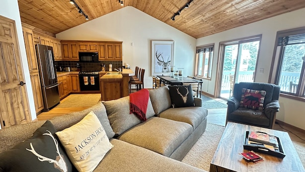 A cozy living room with a large sectional sofa, a coffee table, and a fireplace. The room features wooden ceilings and a kitchen in the background. - Exclusive Teton Pines Home off Teton Village Rd. 5 min from Teton Village Skiing (Wilson)
