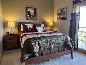 A neatly made bed with a wooden frame, two bedside tables with lamps, a wall-mounted painting, and a window with curtains. - Our Beautiful 3 Bedroom Mountain Retreat to Share with you! (Silverthorne)