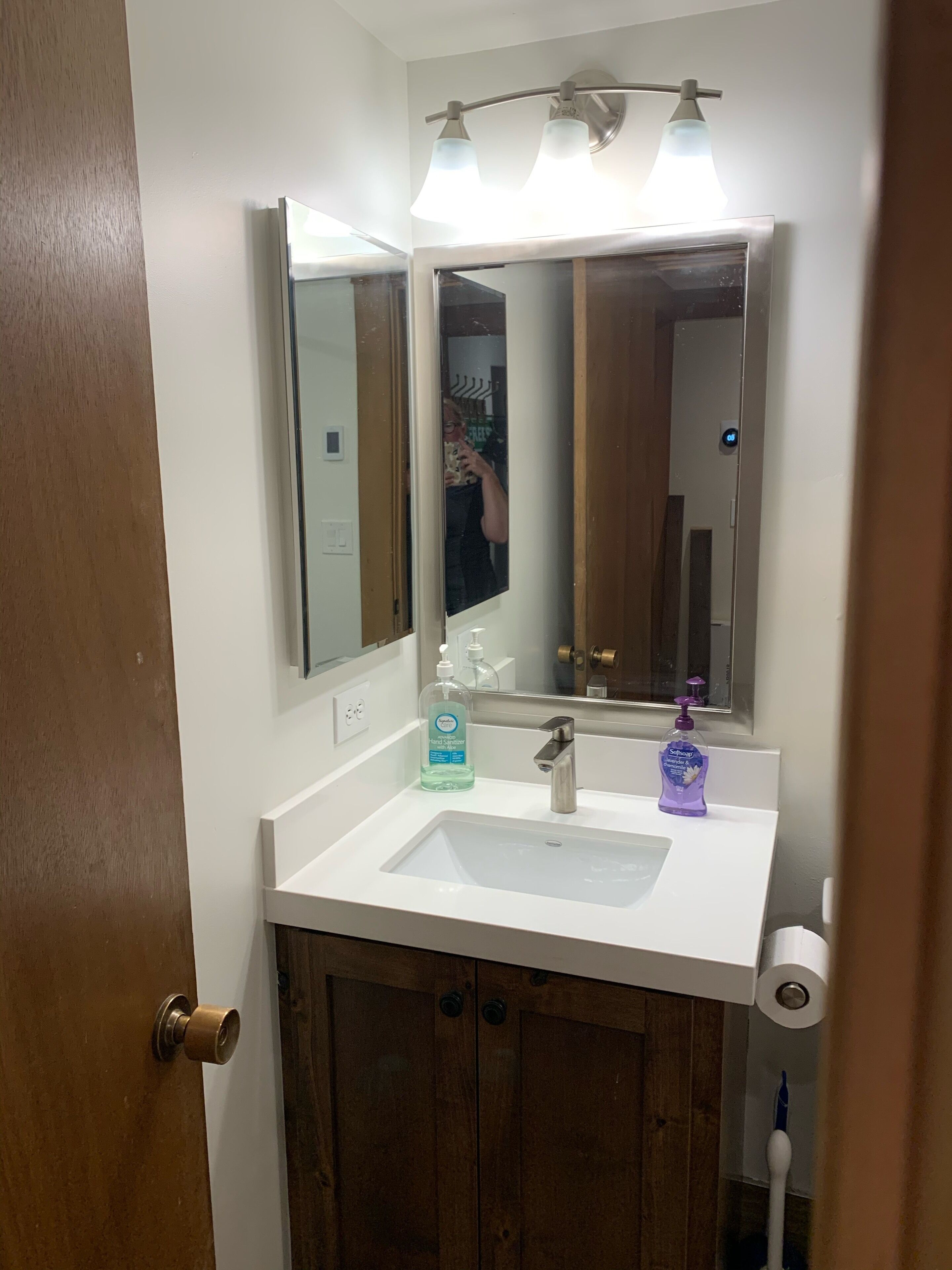 Une salle de bain avec un lavabo blanc, un miroir, un distributeur de savon et un porte-rouleau de papier toilette.