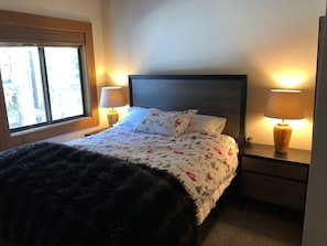 A neatly made bed with floral bedding, two bedside lamps, a wooden headboard, and a window with blinds. - walking distance to skiing and nightlife in Northstar resort (Truckee)