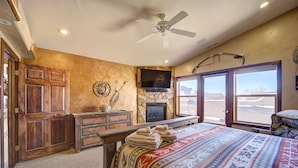A bedroom with a large bed, a television, a ceiling fan, and a stone wall. - High End Southwest Luxury, Spa-like Baths, Steam Shr, Sauna, Garage,Pool,Hot Tub (Moab)