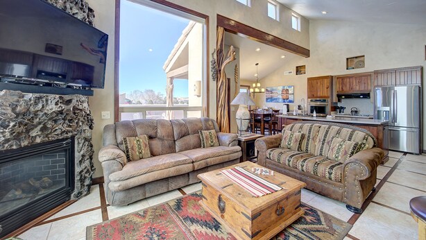 A spacious living room with a fireplace, a large TV, and a view of the kitchen and dining area. - High End Southwest Luxury, Spa-like Baths, Steam Shr, Sauna, Garage,Pool,Hot Tub (Moab)