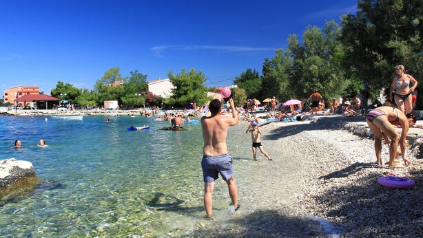 Eine Strandansicht mit klarem Wasser, Personen beim Schwimmen und Sonnenbaden sowie einer felsigen Küstenlinie.