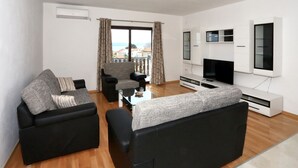 Ein modernes Wohnzimmer mit einer schwarzen Couch, einem grauen Sessel und einem Couchtisch. Ein Fernsehgerät steht auf einer weißen Ablage, und ein Fenster ist mit Vorhängen drapiert.