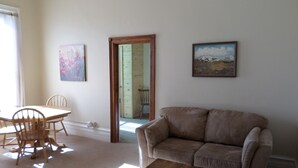 A living room with a beige sofa, a wooden coffee table, and a dining area with a table and chairs. There are two paintings on the wall.