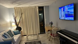 A living room with a sofa, a glass coffee table, a piano, and a television displaying a news program. - HIDDEN GEM 1BR/1BA BEACH PROPERTY AT JETTY EAST, DESTIN , FLORIDA  (Destin)