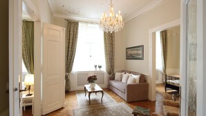 A spacious living room with a wooden parquet floor, a beige sofa, a small table with a vase, and a chandelier.