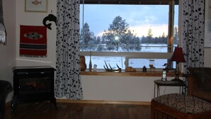 A cozy living room with a fireplace, a window with a view of trees, and a wooden table with a lamp. - Ranch Style Cabin on 20 Acres in the Beautiful Tunk Valley (Riverside)