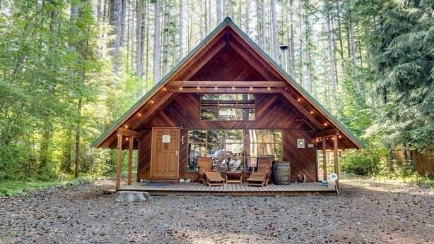 Una cabaña de madera con techo triangular, construida en un bosque. Tiene un porche con asientos y una pequeña mesa.