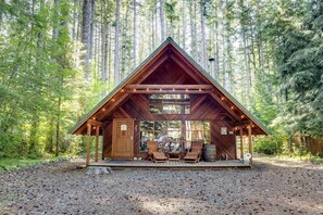 A wooden cabin with a triangular roof, built into a forest. It has a porch with seating and a small table.