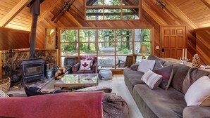 Interior acogedor de una cabaña con chimenea de piedra, un ventanal con vistas al bosque y una zona de estar confortable con sofá y sillones.