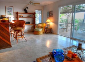 A spacious living area with a dining table, chairs, a bookshelf, and a large window offering a view of the outdoors. - Hale Maluhia - 2 Minutes from the Big Island's Best Beaches (Kamuela)