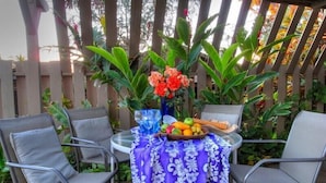 Eine Terrasse mit zwei Metallstühlen, einem Tisch mit violett-blumigem Tischtuch, einer Vase mit orangefarbenen Blumen und einer Obstschale.