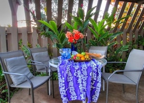 A patio with two metal chairs, a table covered with a purple floral tablecloth, a vase with orange flowers, and a fruit bowl. - Hale Maluhia - 2 Minutes from the Big Island's Best Beaches (Kamuela)