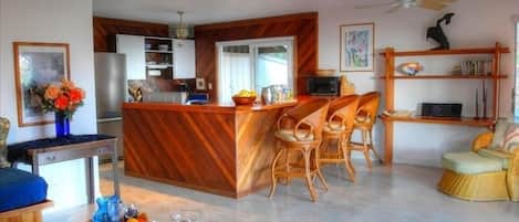 A modern kitchen with a central island, wooden chairs, a dining area with a sofa, and a bookshelf.