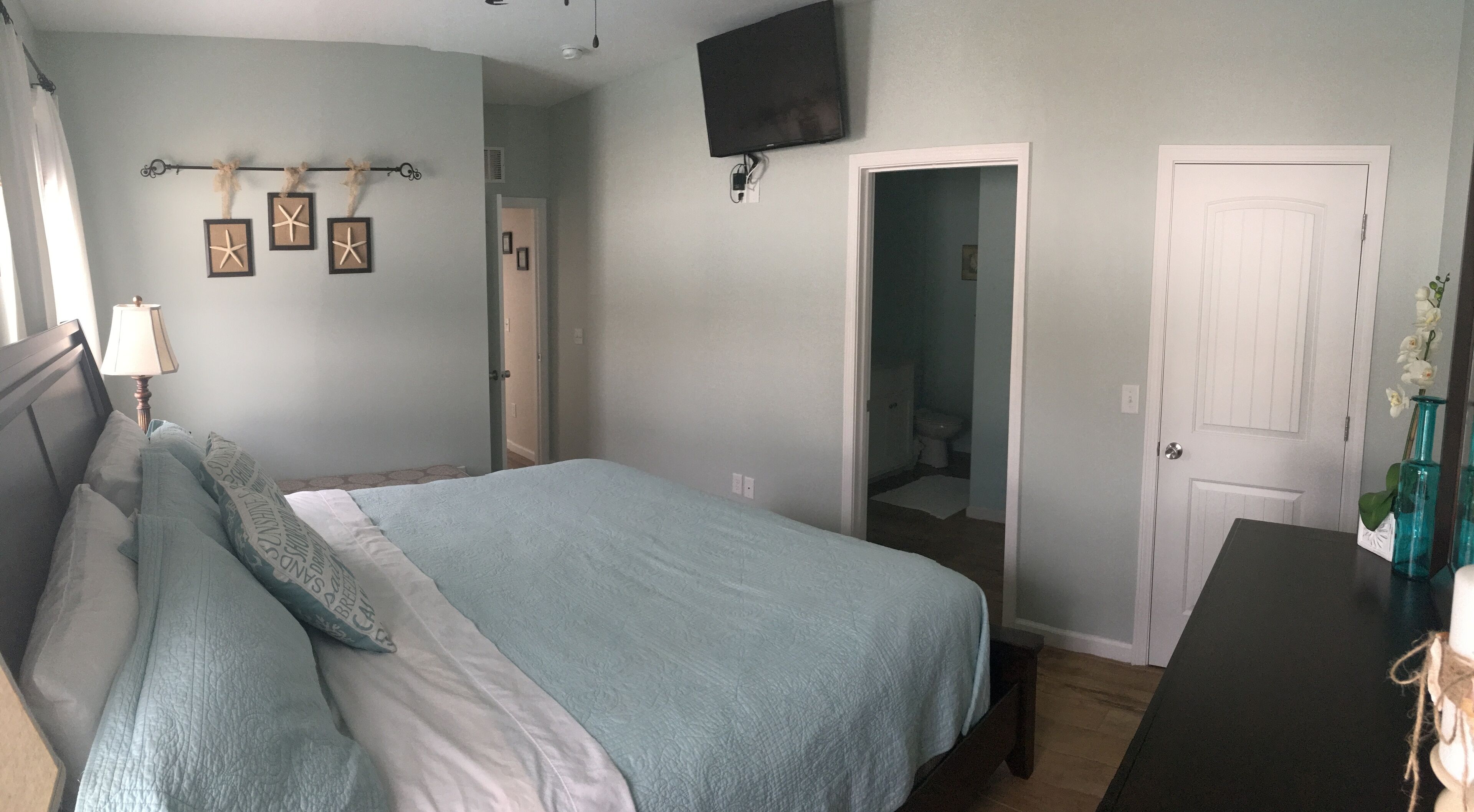 Un dormitorio con cama, cómoda, televisor montado en la pared y una puerta que da a otra habitación.