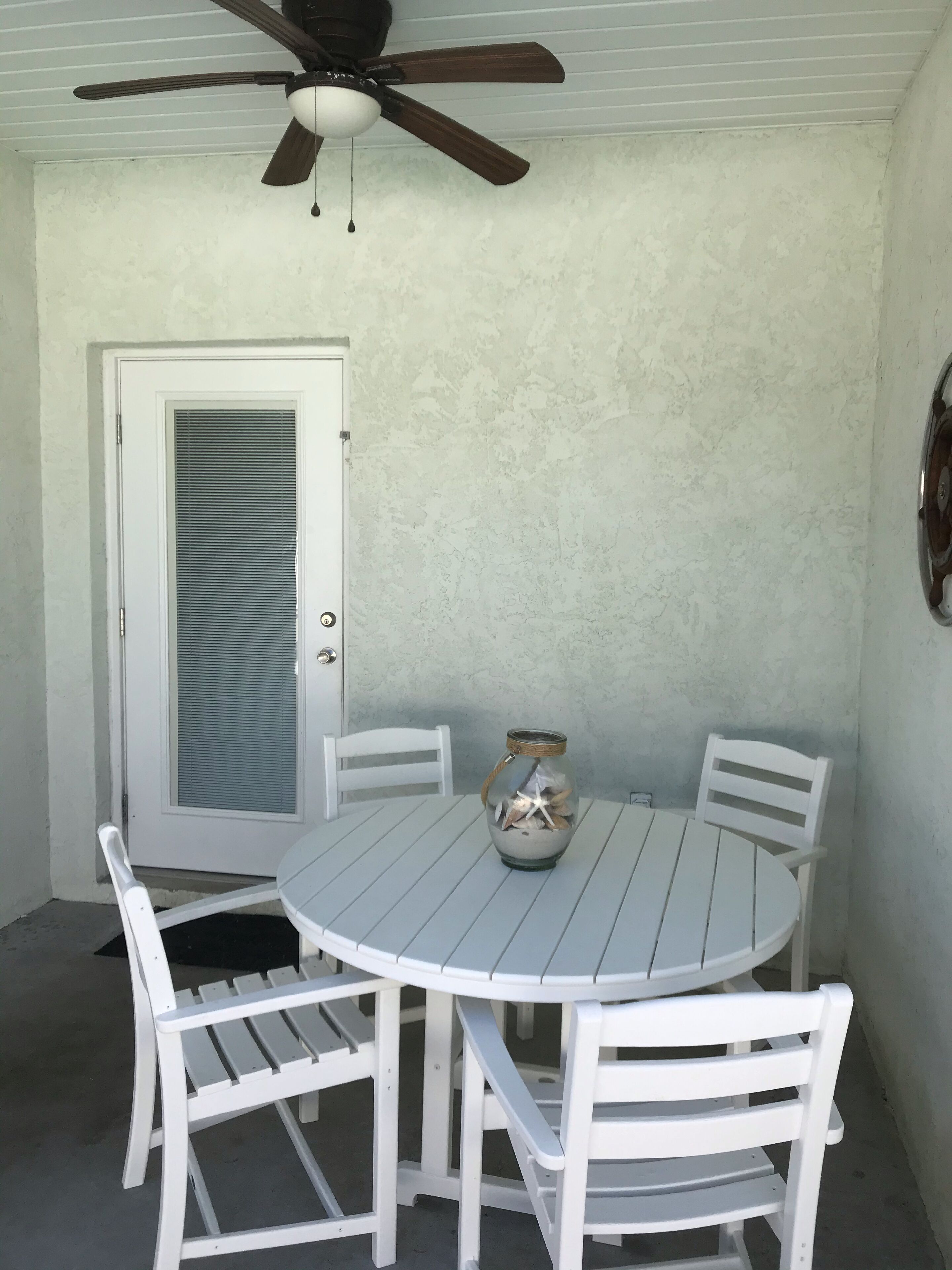 Una mesa redonda blanca con cuatro sillas, un ventilador de techo y una puerta con ventanal.