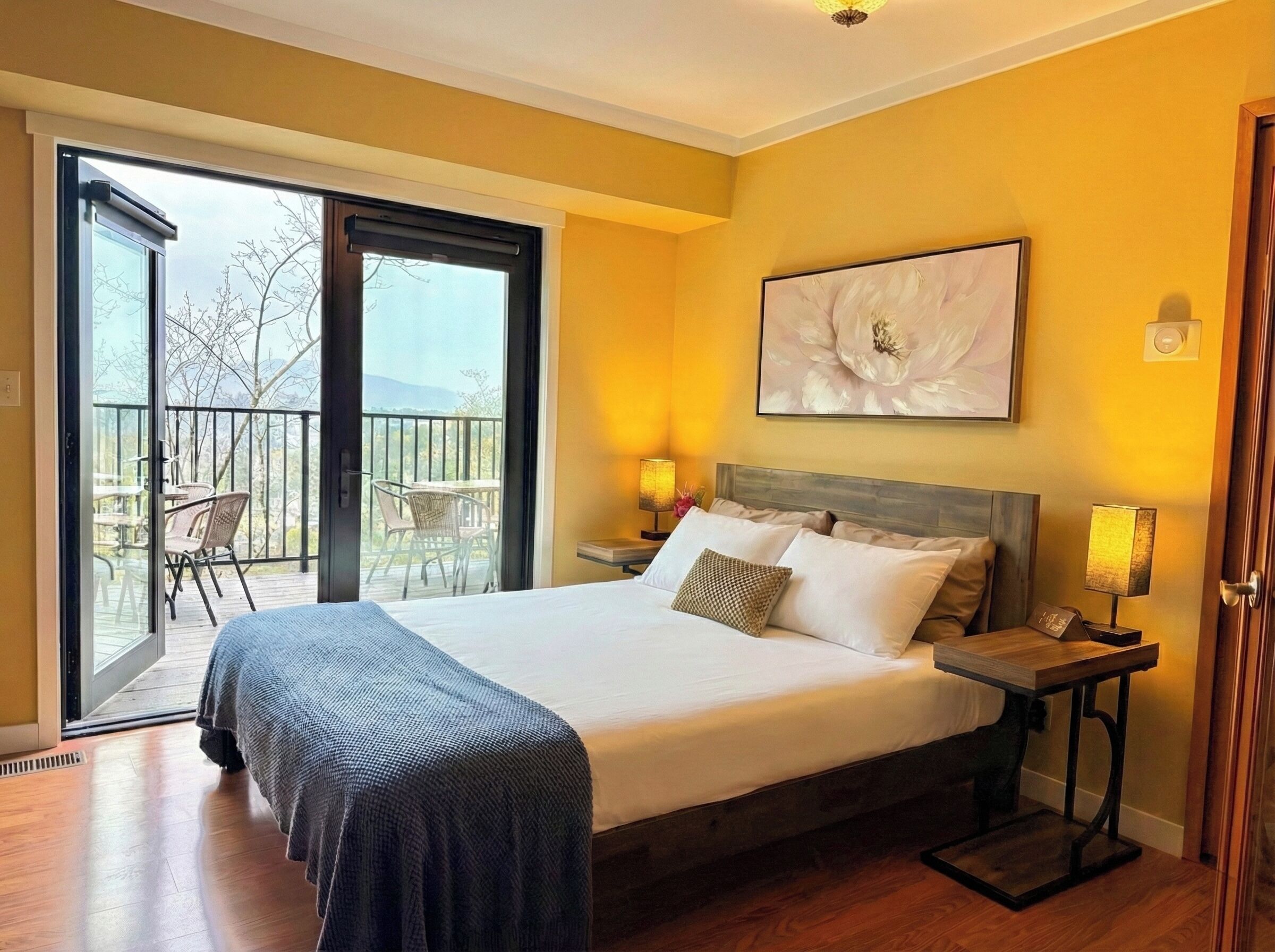 A hotel room with a large bed, two bedside tables with lamps, a balcony with chairs, and a large floral artwork on the wall.